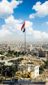 Elevated view of Sana'a cityscape with prominent Yemeni flag under a bright blue sky.