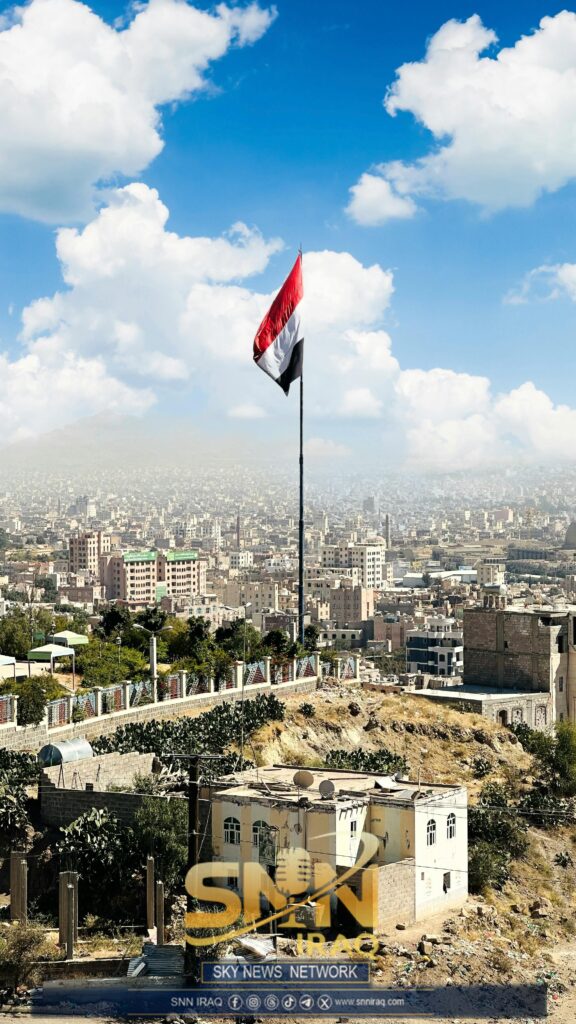 Elevated view of Sana'a cityscape with prominent Yemeni flag under a bright blue sky.